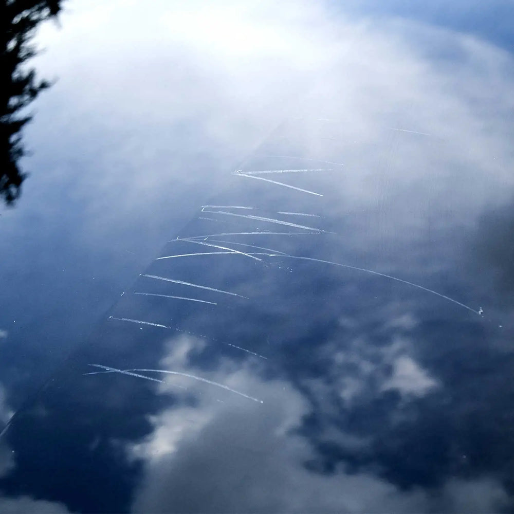 Car paint surface with visible light scratches reflecting sky and clouds before scratch repair