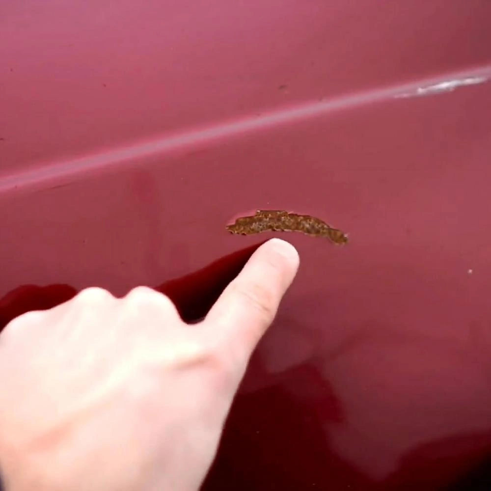 Finger pointing at chipped paint and rust spot on red car surface
