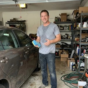 Man standing in garage holding sponge next to dirty car with shelves and tools in background